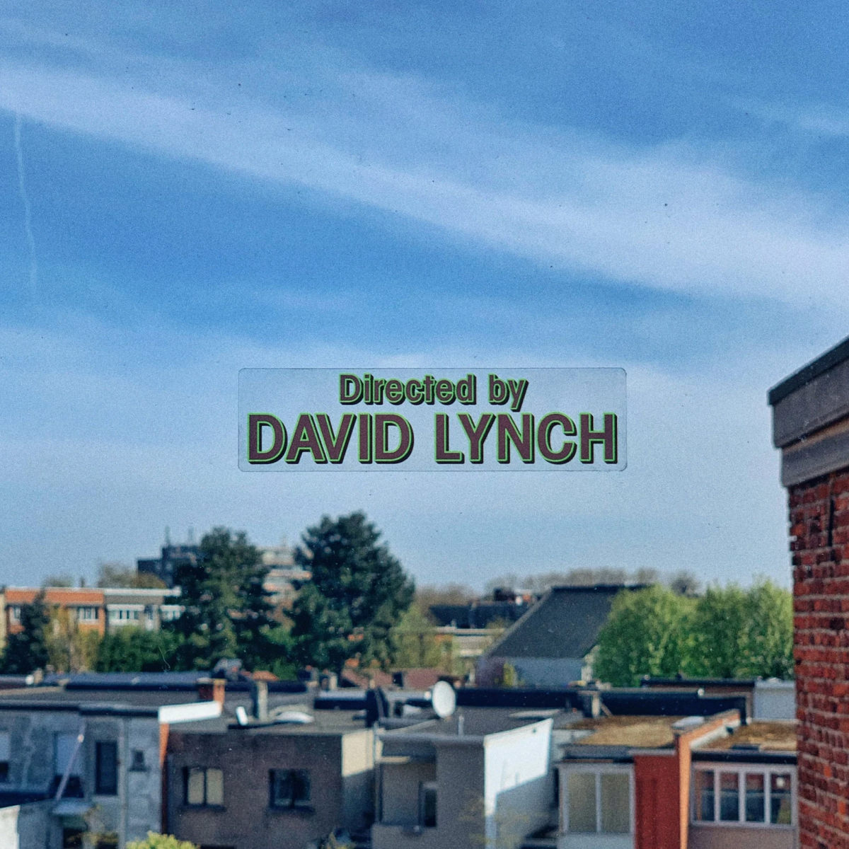 The image shows a suburban neighborhood with houses and trees under a clear blue sky. Superimposed in the middle of the image is text that reads ”Directed by DAVID LYNCH“ in a green, retro-style font, reminiscent of opening or closing credits in a film.