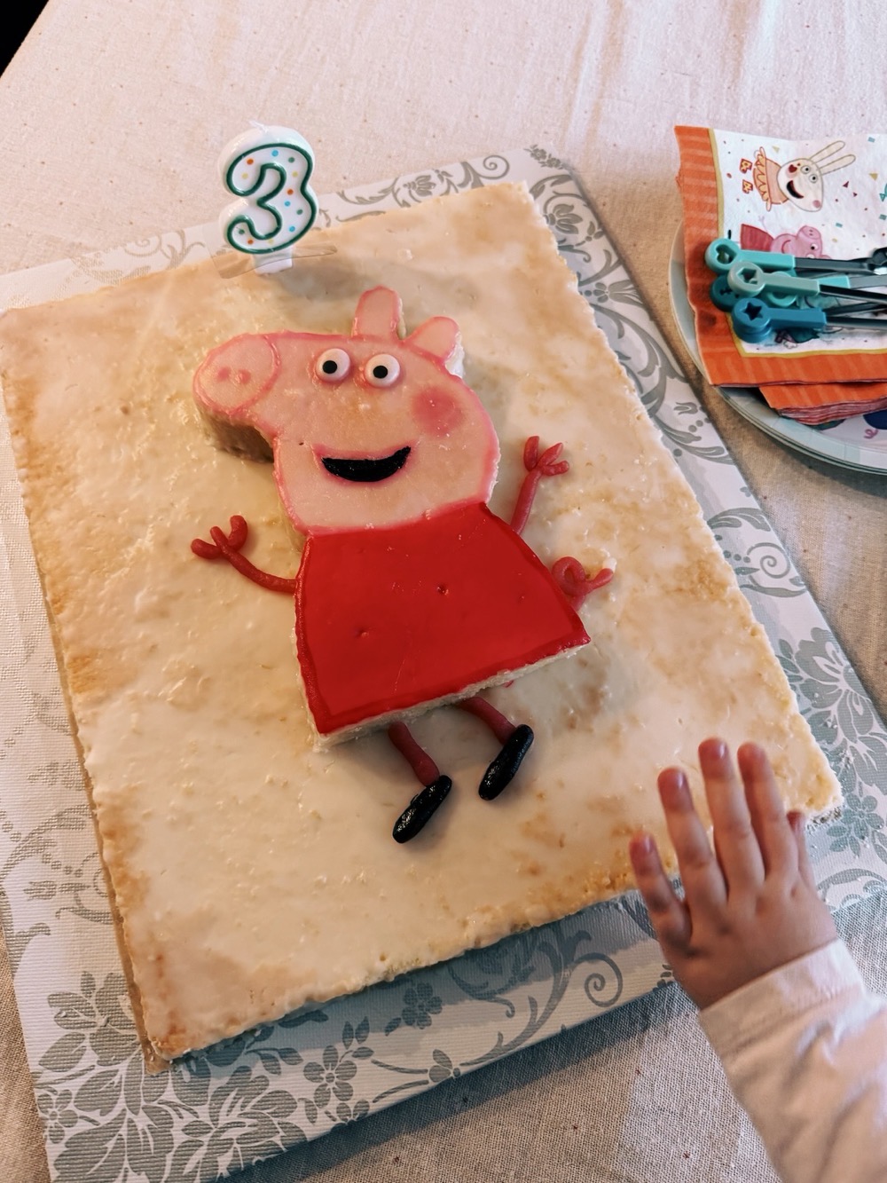 Auf dem Bild ist ein rechteckiger Geburtstagskuchen mit Zuckerguss zu sehen. Darauf liegt eine bunte Figur aus Fondant in Form von Peppa Wutz. Oben auf dem Kuchen steht eine grün-weiß gepunktete Kerze mit der Zahl 3. Rechts im Bild ist die Hand eines kleinen Kindes zu sehen, das offenbar nach dem Kuchen greift. Daneben liegen bunte Servietten und Kuchengabeln.