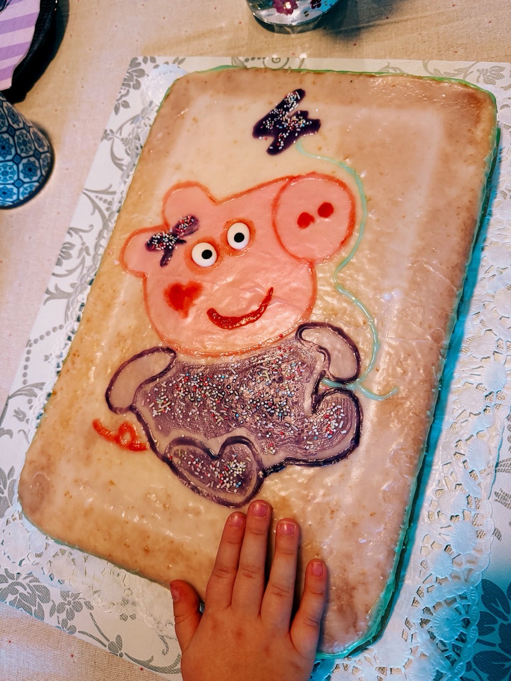 Rechteckiger Geburtstagskuchen mit hellem Zuckerguss und einem Motiv von Baby Evie, der kleinen Schwester von Peppa Wutz. Sie trägt eine Schleife am Ohr und einen lila Strampler mit bunten Zuckerstreuseln und hat ein rotes Herz auf der Wange. Darüber ist eine ebenfalls mit Streuseln verzierte lila Zahl 4 zu sehen, die durch eine grüne Schnur wie ein Luftballon mit der Figur verbunden ist. Am unteren Bildrand greift eine kleine Kinderhand nach dem Kuchen.