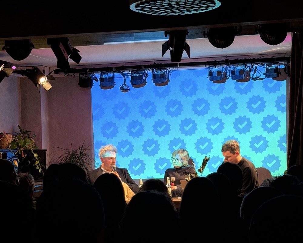 Bühne des Live-Podcasts „Haken dran“ mit drei Personen im Gespräch: Gavin, Franziska und Dennis sitzen nebeneinander, halten Mikrofone und diskutieren vor Publikum. Hinter ihnen leuchtet eine blaue Projektionsfläche mit Haken-dran-Haken, im Vordergrund sind die Silhouetten der Zuhörer*innen zu sehen.