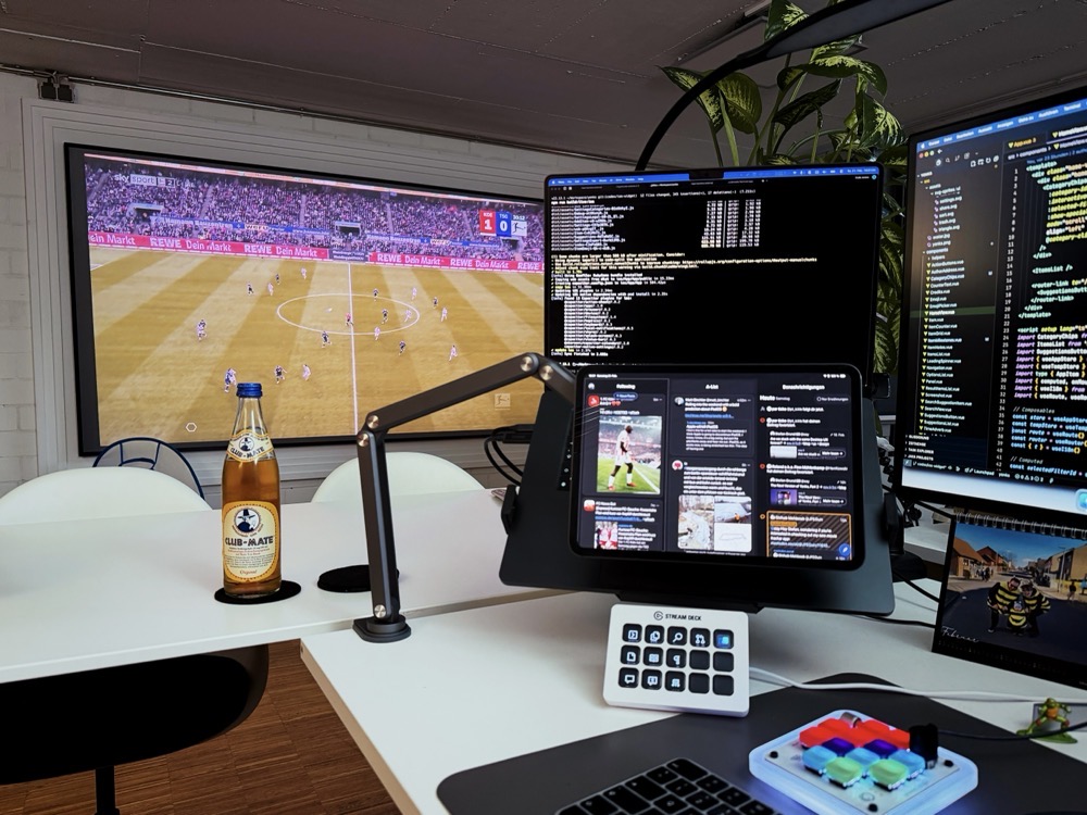 Moderner Arbeitsplatz mit mehreren Monitoren. Auf einer Leinwand im Hintergrund läuft ein Fußballspiel, auf den Monitoren vorne u.a. ein Code-Editor und ein Terminal; davor ein iPad, Stream Deck, beleuchtetes Keypad und eine Flasche Club-Mate auf dem Schreibtisch.