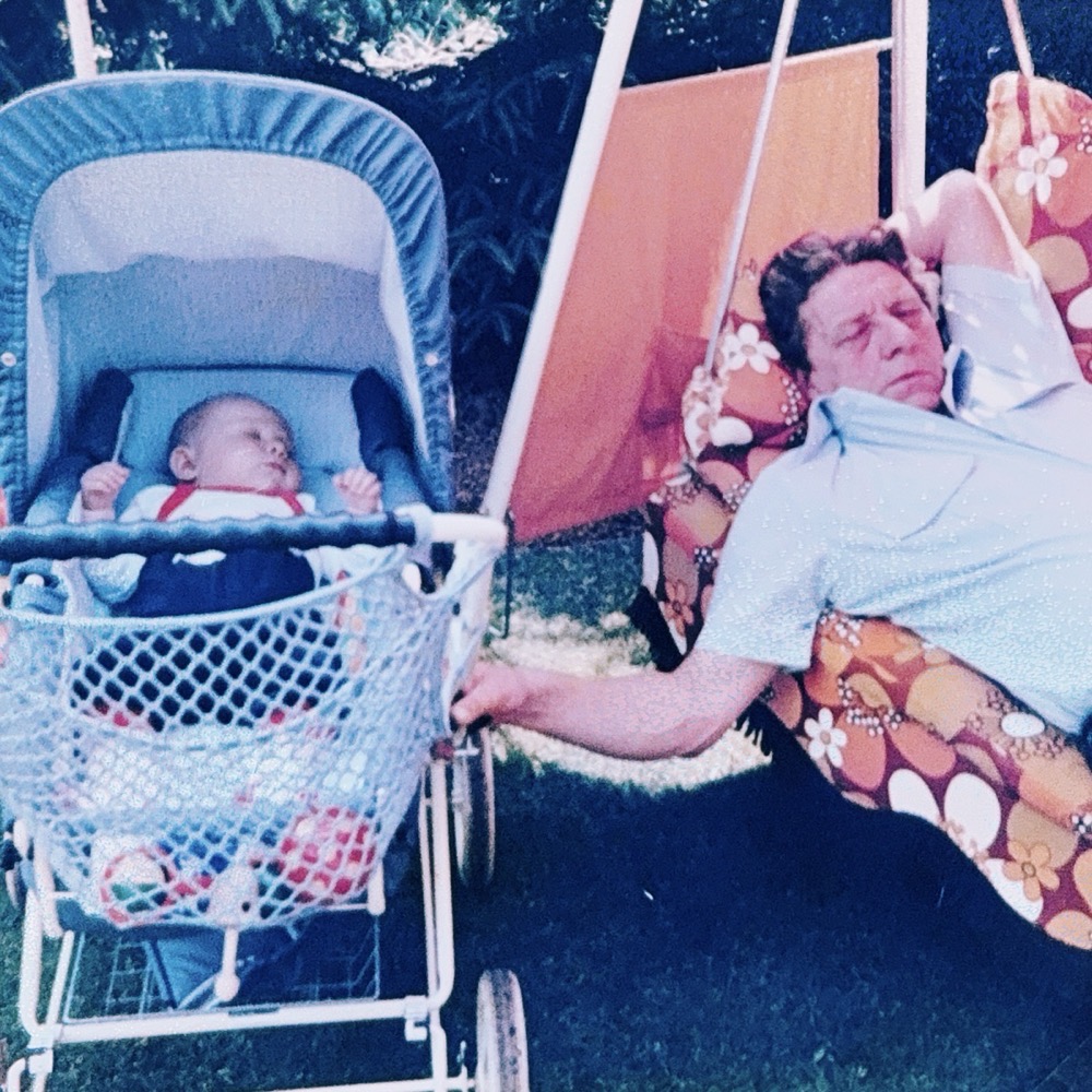 Ein schlafendes Baby liegt in einem Kinderwagen neben seinem schlafenden Großvater auf einer Hollywoodschaukel im Garten. Der Großvater hält den Kinderwagen mit einer Hand fest.